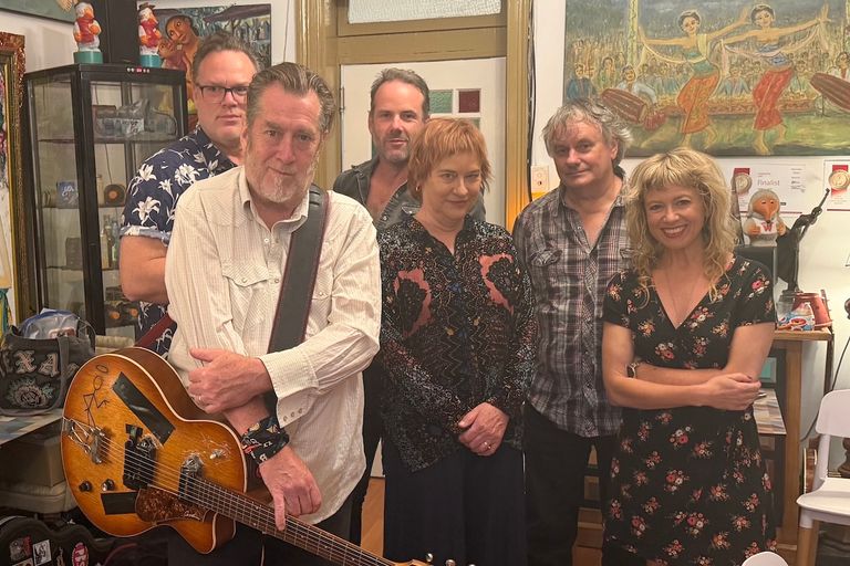 Alt text (144 characters): Australian band Mick Thomas' Roving Commission posing together indoors, with frontman Mick Thomas holding a vintage acoustic-electric guitar.